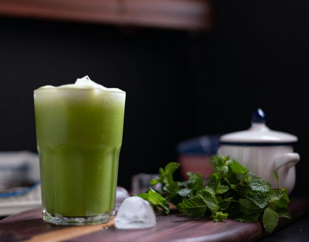 Copo de suco verde ao lado de ingredientes frescos como limão, gengibre e folhas verdes